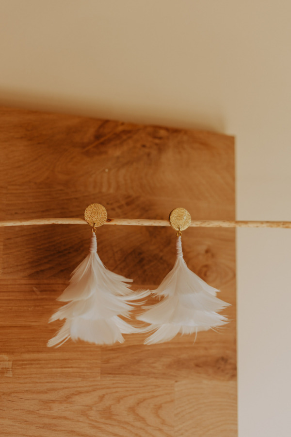 boucles d'oreilles blanche tout en volume en plume d'oie, les boucles sont présenter devant une planche de bois, elles sont idéale pour un mariage. On utilise l'art de la plumasserie