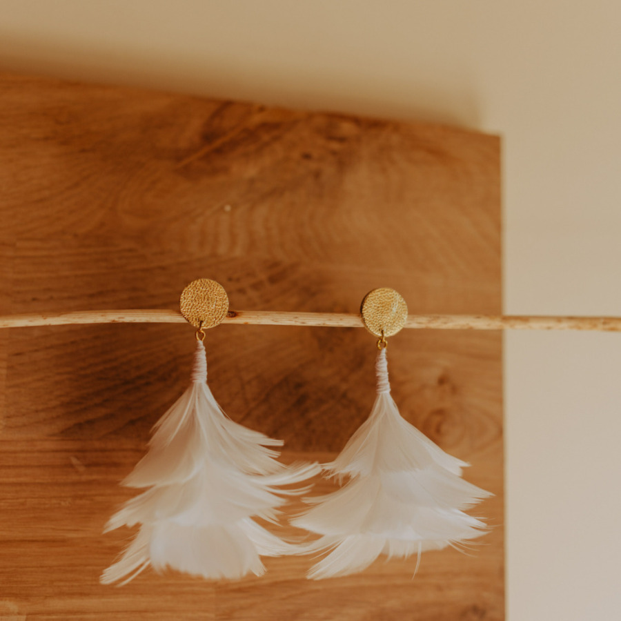 boucles d'oreilles blanche tout en volume en plume d'oie, les boucles sont présenter devant une planche de bois, elles sont idéale pour un mariage. On utilise l'art de la plumasserie